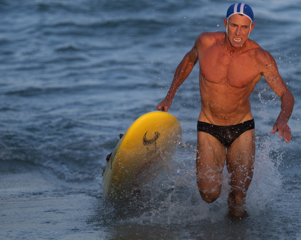 A surf lifesaving competitor runs out of the ocean carrying a yellow rescue board during a beach race. Water splashes around his legs as warm low-angle sunlight highlights his face and upper body against the blue water in the background.