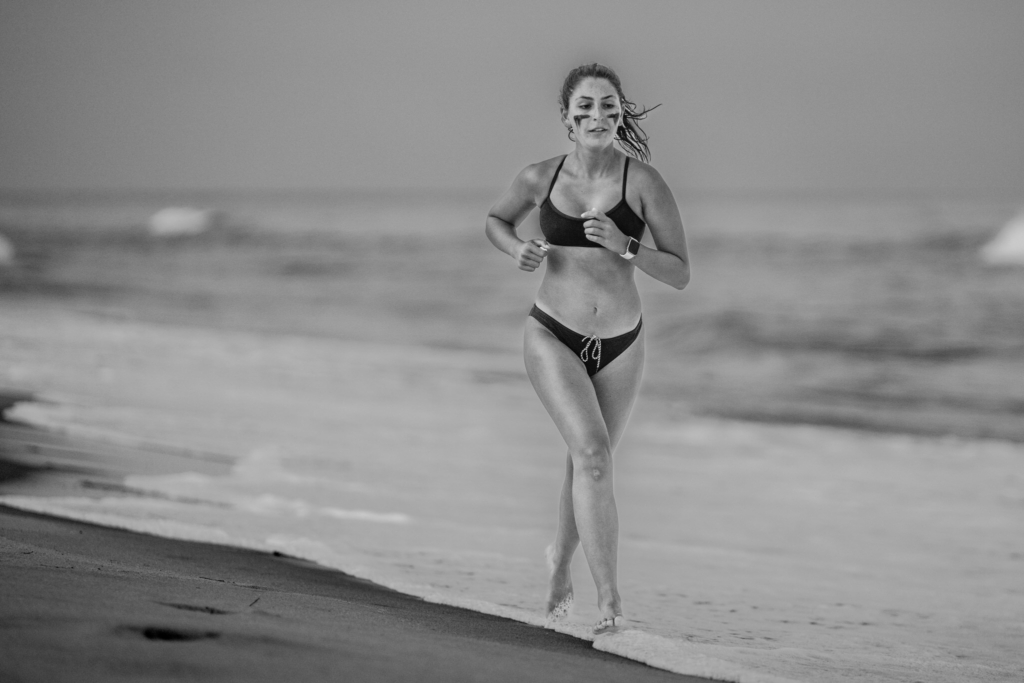 A female surf lifesaving athlete runs along the water’s edge after a swim leg, wearing goggles and a racing swimsuit. The black-and-white image captures water droplets on her skin and the textured shoreline fading into the background.