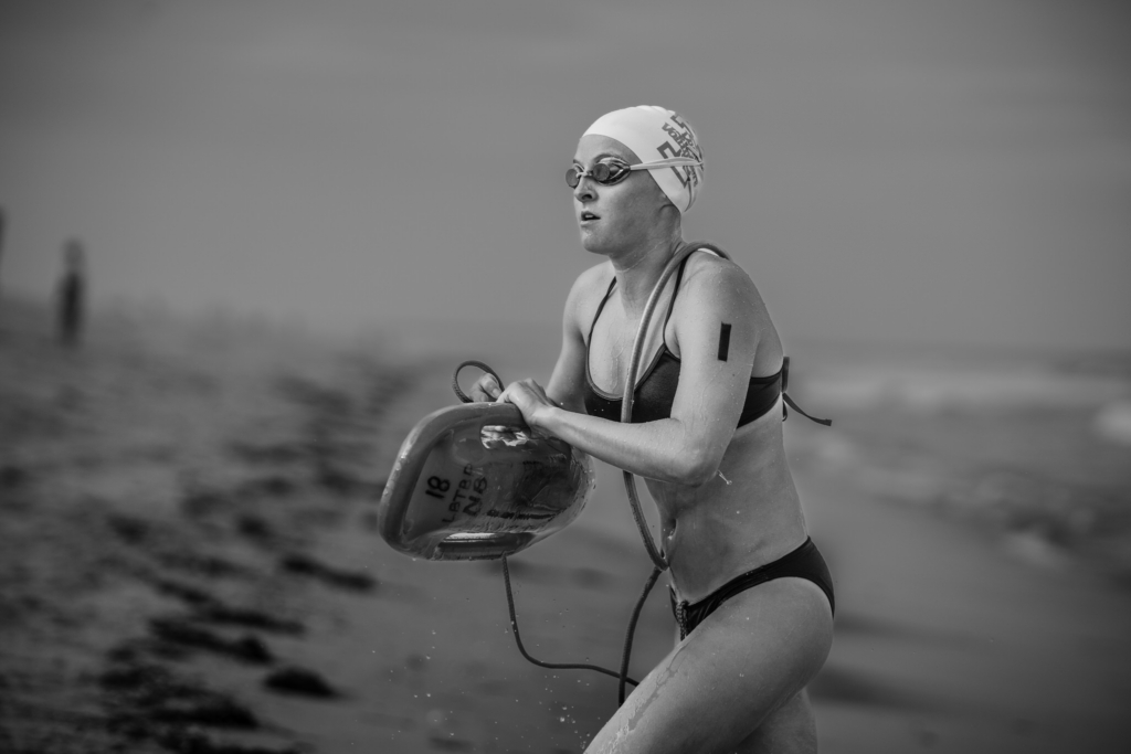 A female surf lifesaving competitor emerges from the ocean carrying a rescue tube during a race. Wearing a swim cap and goggles, she moves through shallow water with the beach and surf softly blurred behind her in this black-and-white photograph.