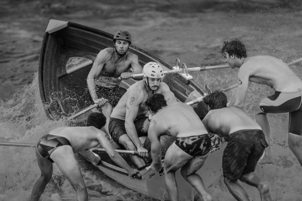 A surf boat rescue team battles through breaking waves as crew members push and stabilize the boat in the shoreline surf. The black-and-white image captures splashing water, helmets, and the coordinated effort of the athletes during a surf lifesaving