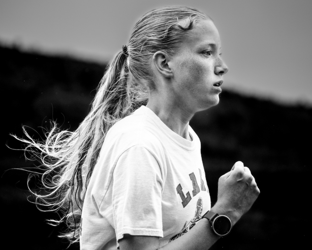 A runner is captured in a close-up black-and-white portrait during training, her ponytail moving behind her as she runs. Soft light highlights her face and expression, while the dark, blurred background isolates her against the landscape.