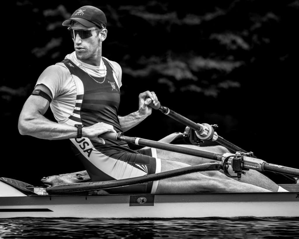 A sculling athlete rows a single racing shell across calm water, captured in a close-up black-and-white portrait. Wearing sunglasses and a USA racing uniform, he turns his head to the side while holding the oars, with a dark, blurred shoreline behind him.