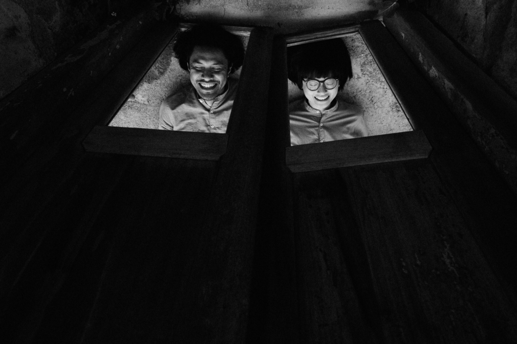 A black-and-white photograph shows two people lying side by side beneath wooden panels, viewed from above through narrow openings. Soft light illuminates their smiling faces against the dark surrounding structure, creating an intimate and playful atmosphere with strong contrasts of light and shadow.