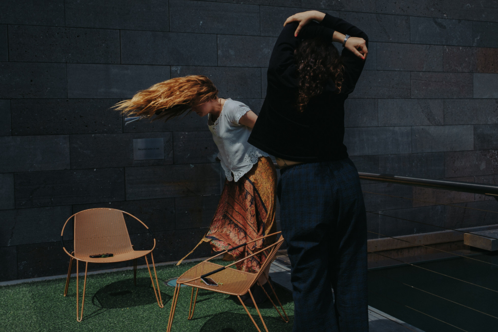 Two people move expressively in an outdoor courtyard beside a dark stone wall, surrounded by metal chairs on a patch of artificial grass. One person bends forward with long hair sweeping through the air, while the other stretches their arms overhead, creating a dynamic sense of motion under directional natural light.
