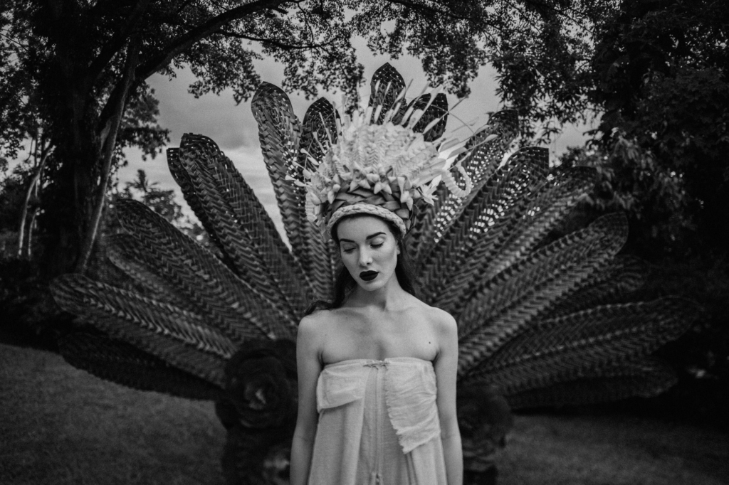 A black-and-white portrait shows a woman standing outdoors wearing an elaborate woven headdress with large fan-like elements extending behind her. Soft natural light illuminates her face and shoulders as she looks downward, while dark trees and foliage create a dramatic backdrop that emphasizes the intricate textures of the headpiece.