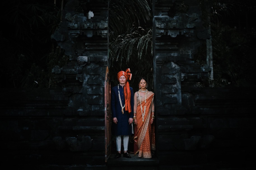 A couple wearing traditional South Asian wedding attire stands side by side in the entrance of a dark stone temple structure. Soft, focused light illuminates their richly detailed clothing and jewelry, while the surrounding architecture and deep shadows create a calm, ceremonial atmosphere.
