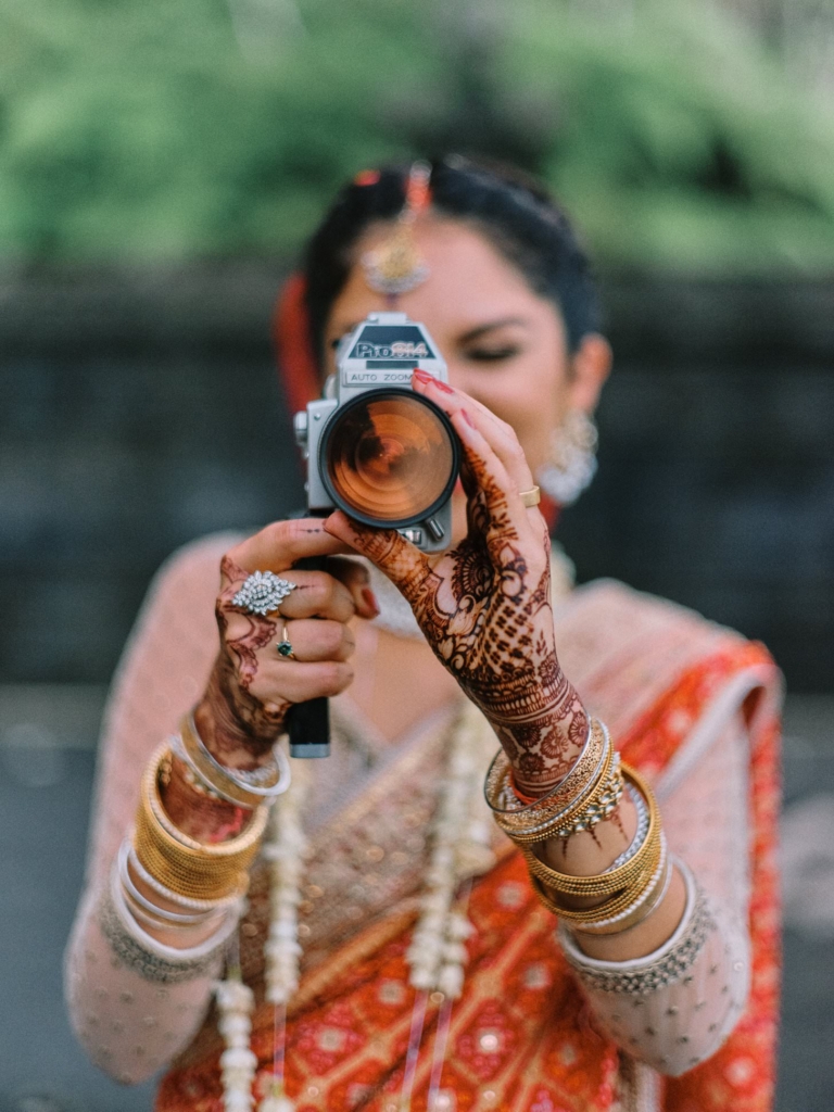A woman dressed in traditional South Asian wedding attire holds a vintage camera up to her face as if taking a photograph. Her hands are decorated with intricate henna patterns and layered gold jewelry, while soft natural light and a blurred background draw attention to the camera and her detailed attire.