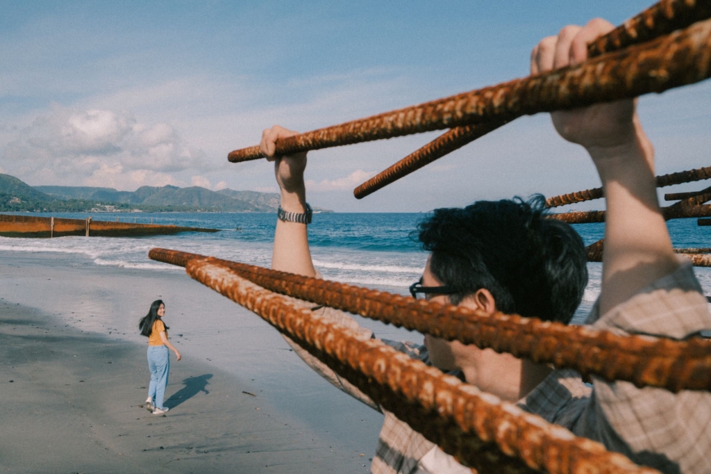 A person stands on a sandy beach holding onto several rusted metal bars that extend diagonally across the foreground, partially framing the scene. In the distance, another person walks along the shoreline and looks back toward the camera, while gentle waves, mountains, and a bright blue sky create a relaxed coastal atmosphere.