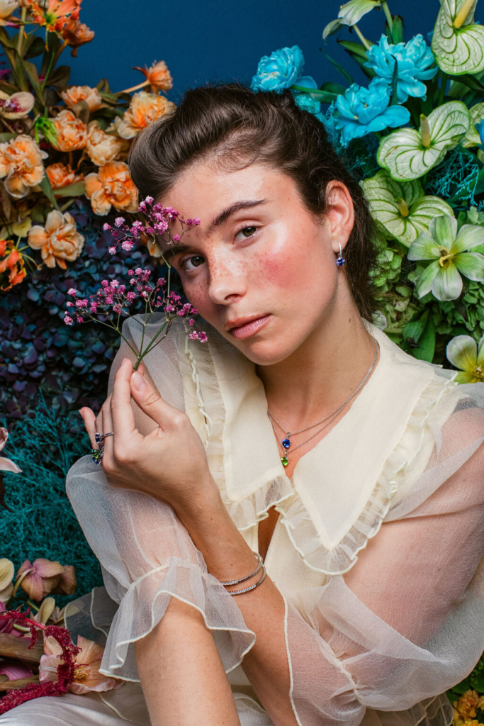 A young woman sits among a dense arrangement of colorful flowers, holding a small sprig of pink blossoms near her face. She wears a sheer cream blouse with ruffled details and delicate jewelry, while soft, even light highlights her features against a vivid backdrop of orange, blue, green, and white blooms, creating a rich and intimate portrait.
