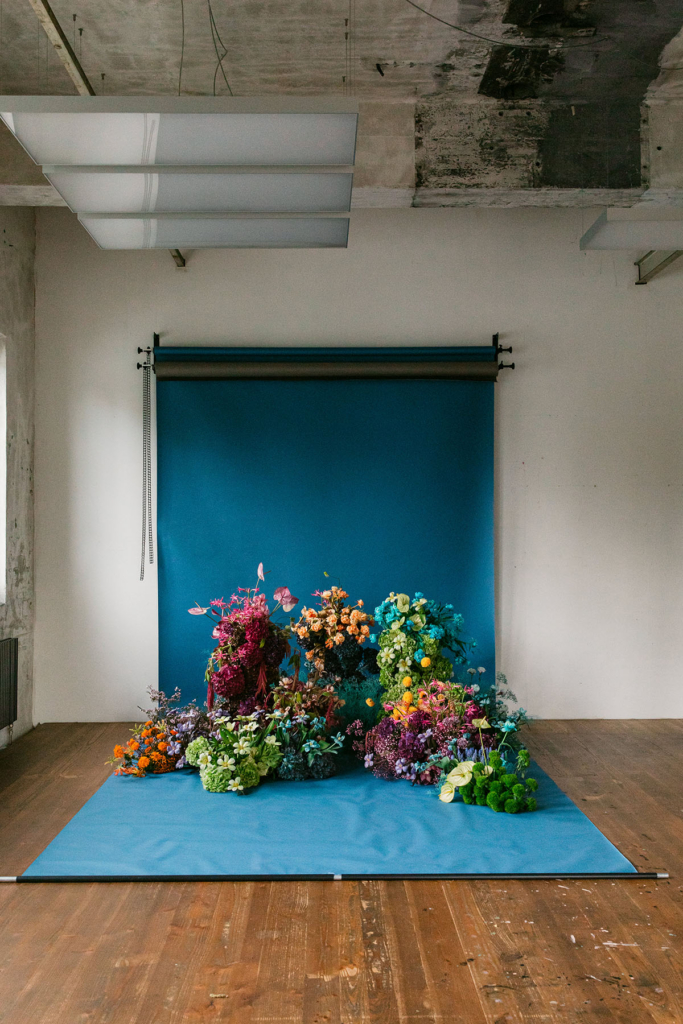 A vibrant floral installation sits on a blue backdrop inside an industrial-style studio with wooden floors and exposed concrete ceilings. Clusters of colorful flowers in pink, orange, green, blue, and purple spill across the floor, illuminated by soft overhead light that contrasts the raw architectural space with the lush arrangement.