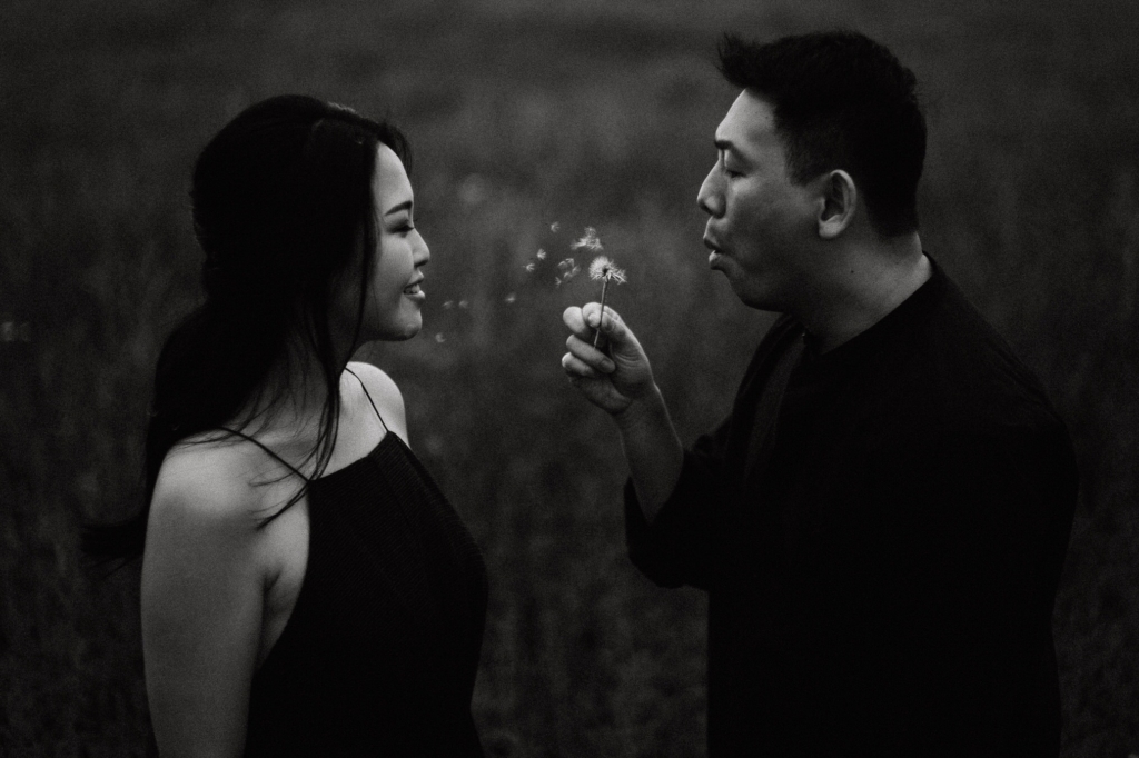 A black-and-white portrait shows two people facing each other in an open field, both dressed in dark clothing. One person holds a dandelion and blows its seeds toward the other, who smiles softly as the seeds drift between them, creating a playful and intimate moment against a blurred natural background.