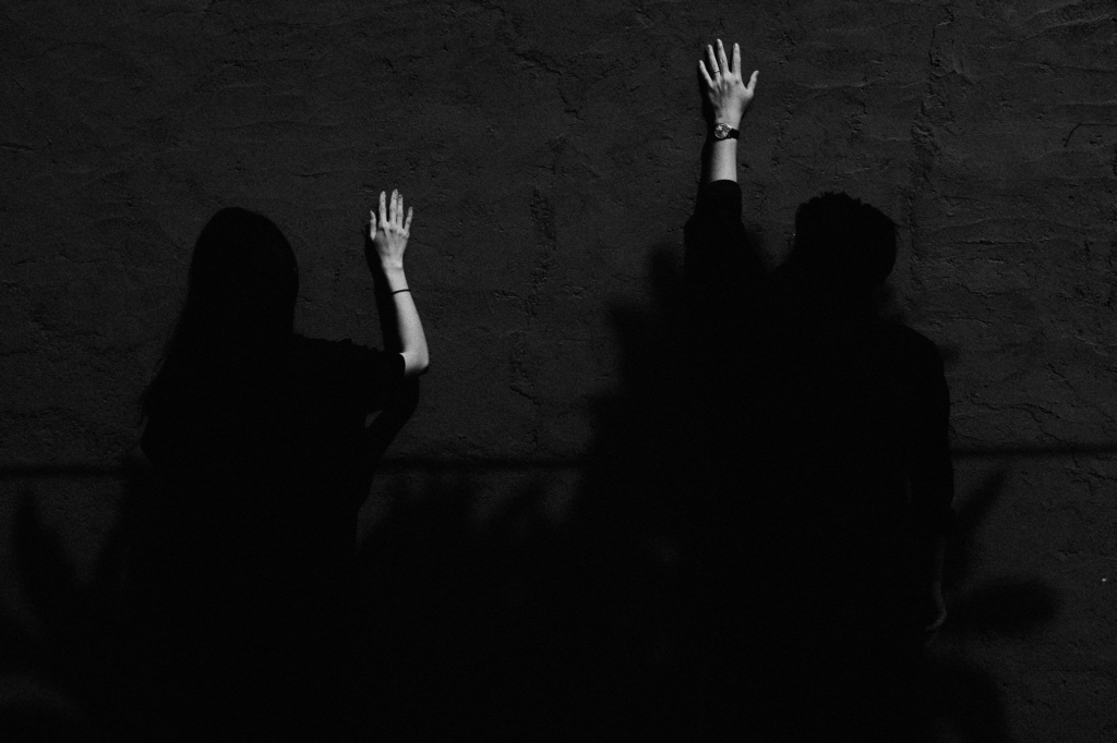 A black-and-white photograph shows two people standing against a textured wall, their bodies mostly in shadow while their raised hands are illuminated by a narrow beam of light. The strong contrast and simple composition create a quiet, dramatic atmosphere, drawing attention to the gesture and the interplay between light and darkness.