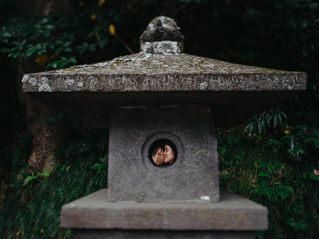A weathered stone lantern stands in the foreground of a lush garden, surrounded by dark green foliage and soft natural light. Through a small circular opening in the lantern, a couple is visible sharing a kiss, creating an intimate focal point framed by the textured stone structure.