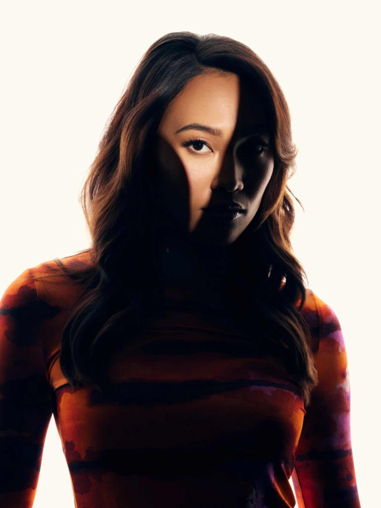 A woman with long dark hair faces the camera in a close-up portrait, wearing a patterned long-sleeve top. Dramatic lighting leaves half of her face in shadow while the other half is brightly lit, creating a bold, high-contrast look against a plain light background.