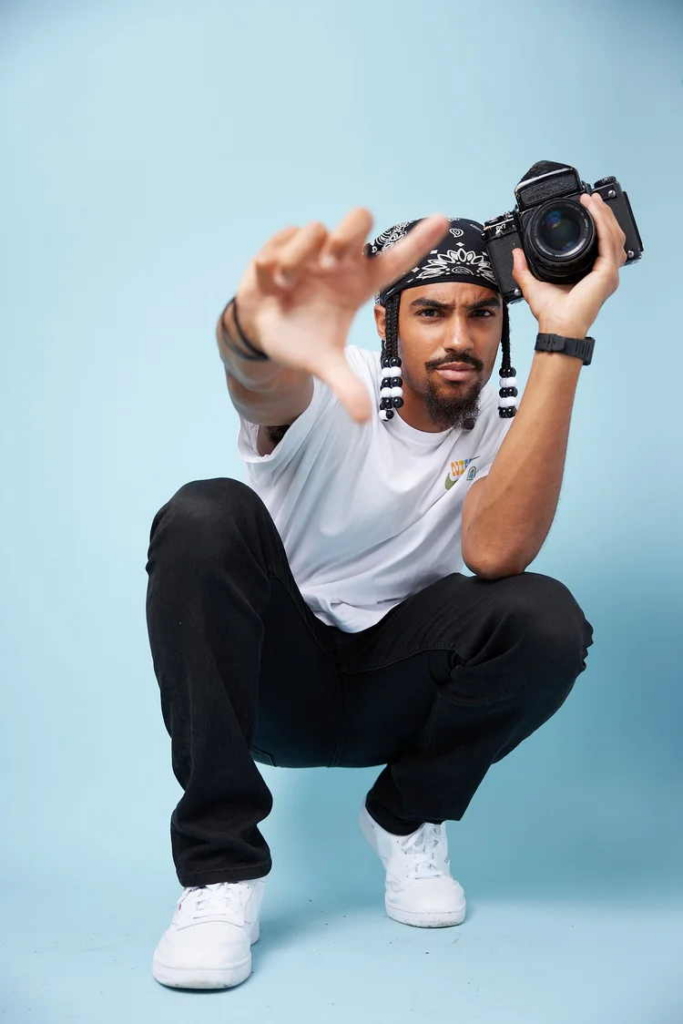 A photographer crouches against a light blue background, holding a camera up beside his face while reaching one hand toward the camera. He wears a white T-shirt, black pants, white sneakers, and a patterned bandana.