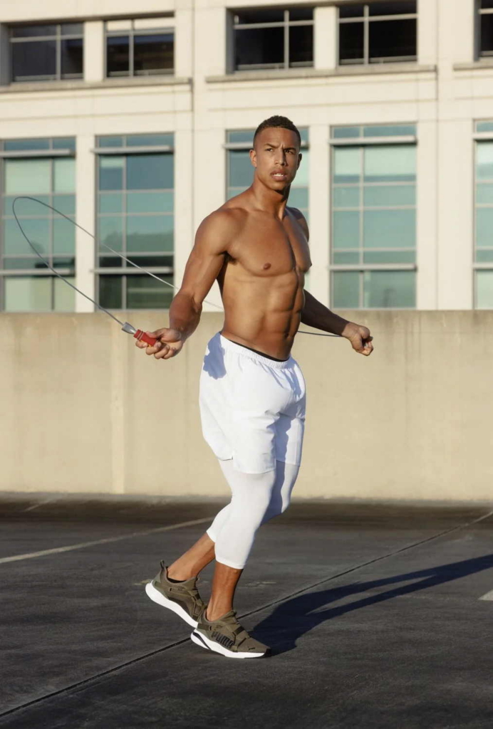 A shirtless man wearing white athletic leggings and sneakers jumps rope on an outdoor rooftop in bright sunlight. A modern building stands in the background as he looks to the side in mid-step.