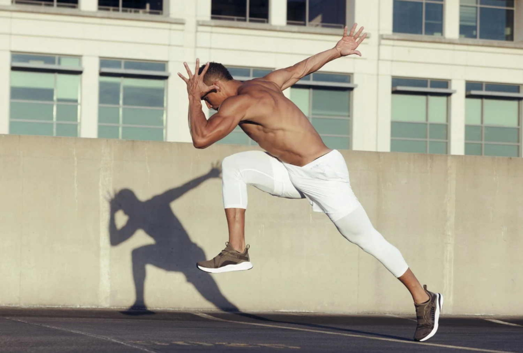 A shirtless man wearing white athletic leggings and dark sneakers is captured mid-jump with his arms outstretched in an outdoor urban setting. Bright sunlight casts a sharp shadow of his pose onto a concrete wall, with a modern building visible in the background.