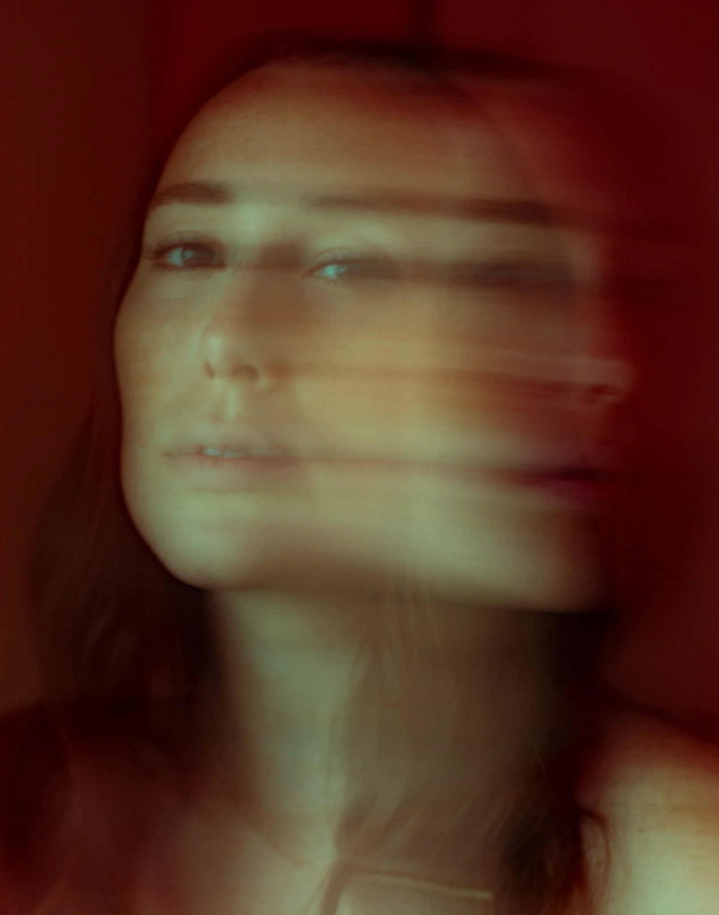 A close-up portrait of a woman with long dark hair is shown with intentional motion blur creating a layered effect across her face. Warm red and orange tones fill the background, while one eye and part of her face remain more clearly visible.