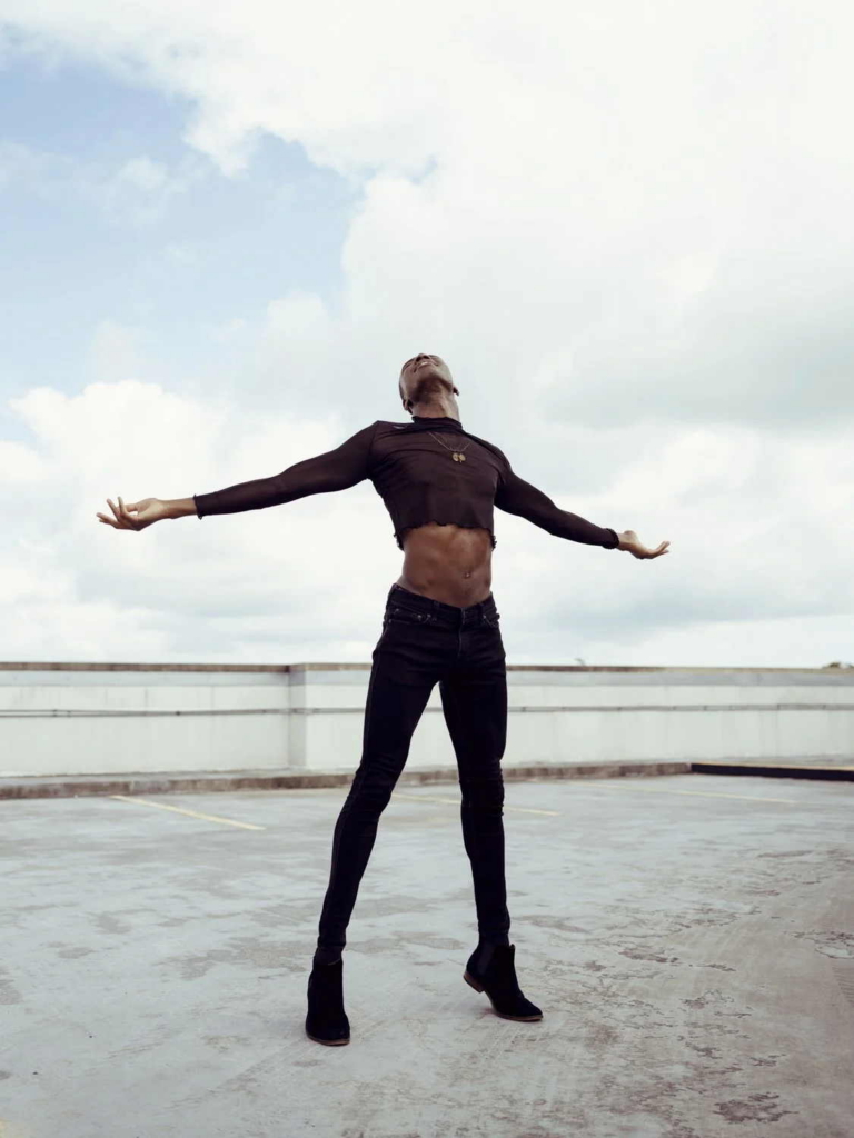 A person stands on an open rooftop with their arms stretched wide and their face turned toward the sky. They wear a black long-sleeved top, black pants, and black boots, with a cloudy sky filling the background.