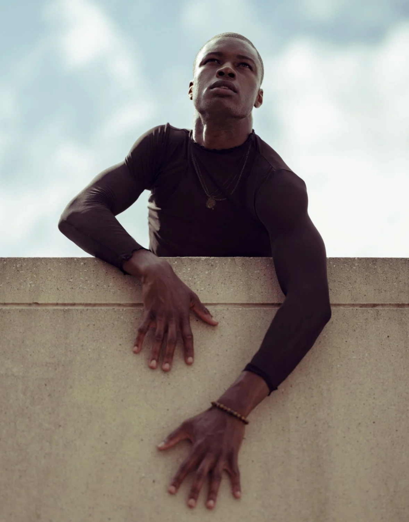 A person in a fitted black long-sleeved shirt leans over a concrete wall, looking upward toward the sky. One arm rests on the top of the wall while the other hangs down, with soft daylight and clouds in the background.