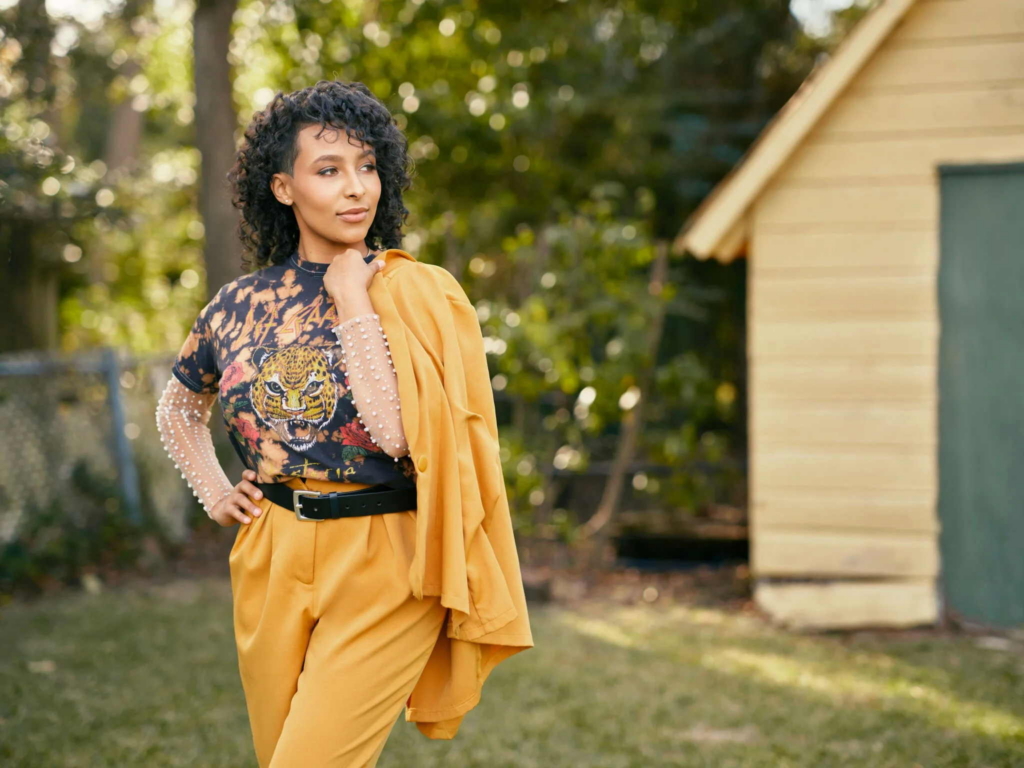 A woman stands outdoors near a small wooden building, looking off to the side with one hand on her hip. She wears mustard-colored pants and a matching jacket over one shoulder, paired with a graphic shirt, while sunlight softly lights the trees in the background.