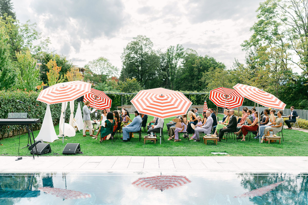 Guests sit on a lawn during an outdoor wedding ceremony beside a swimming pool, shaded by large red-and-white striped umbrellas. The scene is bright and colorful, with greenery surrounding the gathering and reflections of the umbrellas visible in the water.