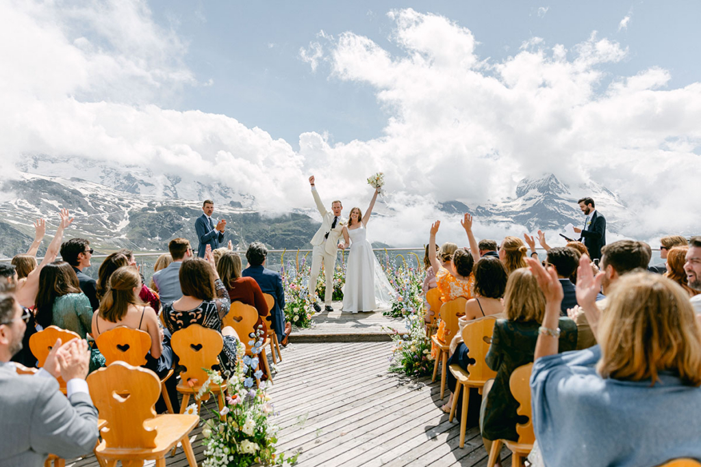 An outdoor wedding ceremony takes place on a wooden terrace surrounded by guests, with the newly married couple standing at the front and raising their arms in celebration. Snow-capped mountains and dramatic clouds fill the background, while guests applaud and lift their hands in a bright, festive atmosphere.