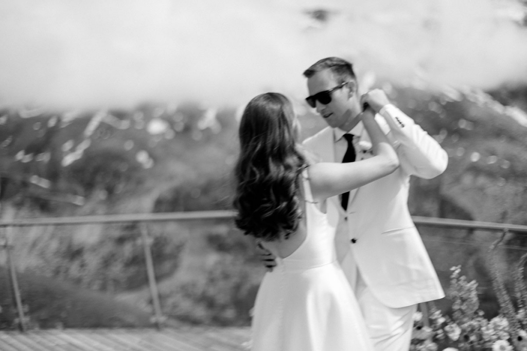 A bride and groom dance together outdoors on a terrace overlooking distant mountains, captured in black and white with soft motion blur. The groom wears a light-colored suit and sunglasses, while the bride’s flowing dress and raised arms add a sense of movement and energy to the scene.