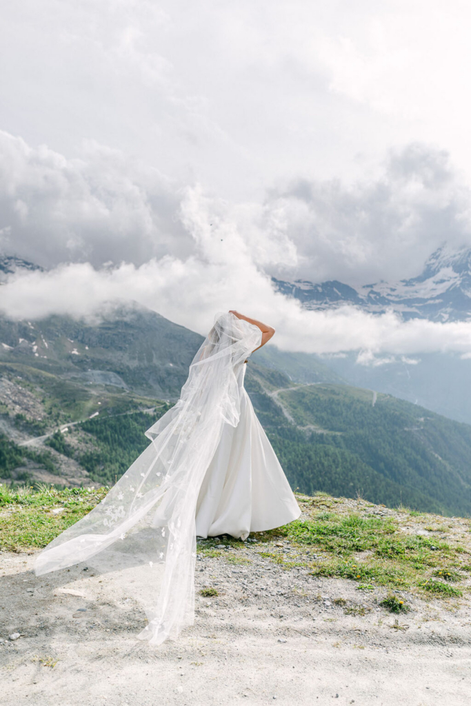 A bride stands on a grassy hillside overlooking a lake and distant mountains, holding out her long veil as it catches the breeze. Soft daylight and layered clouds create an airy, peaceful setting, with the landscape stretching into the distance behind her.