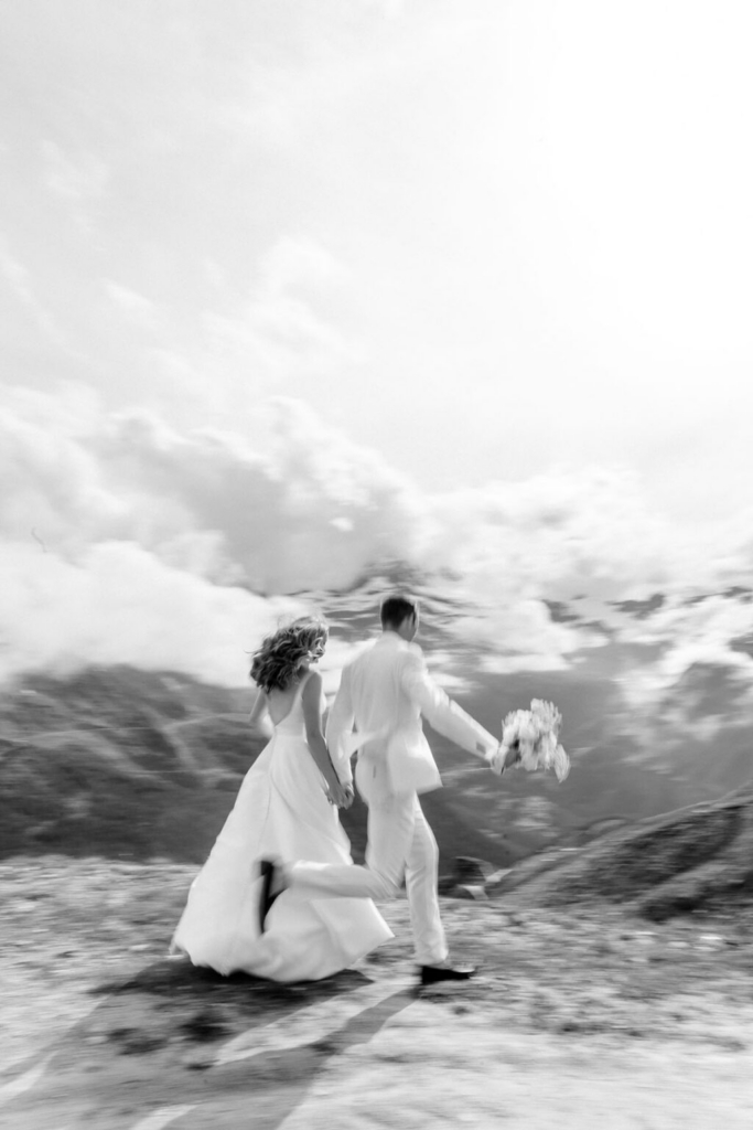 A bride and groom run hand in hand across an open mountain landscape, photographed in black and white with motion blur that emphasizes their movement. Snow-covered peaks and dramatic clouds fill the background, creating a dynamic and expansive setting.