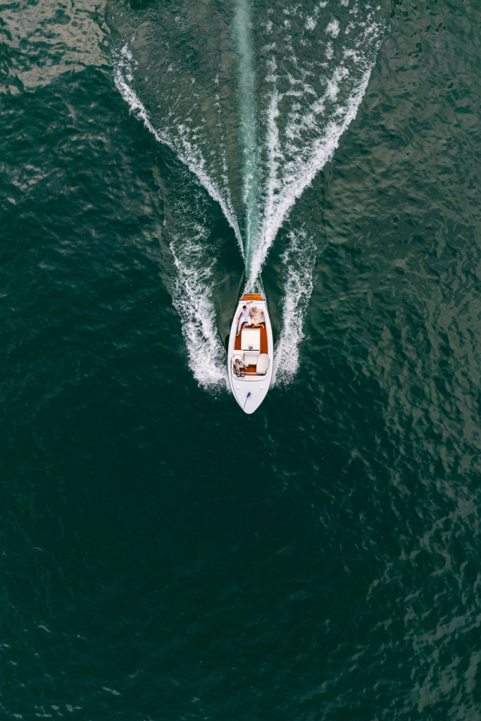 An aerial view shows a small motorboat traveling across deep blue water, leaving a symmetrical white wake behind it. Several people sit inside the boat as it moves through the lake, creating strong geometric patterns on the water’s surface.