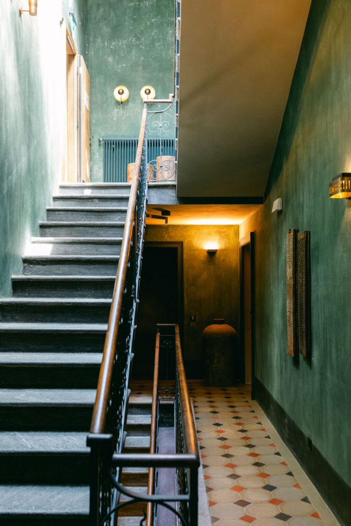 An interior staircase with dark stone steps and ornate railings rises through a narrow hallway with textured green walls. Warm wall sconces illuminate the space, highlighting patterned floor tiles, wooden doors, and a large ceramic vessel beneath the stairs, creating a quiet and inviting atmosphere.