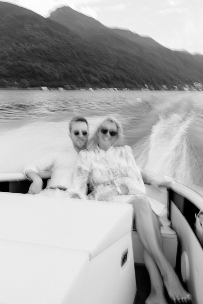 A couple sits together on a motorboat crossing a lake, photographed with intentional motion blur as water trails behind them. The black-and-white image combines the movement of the boat with the surrounding mountains and shoreline in the distance.
