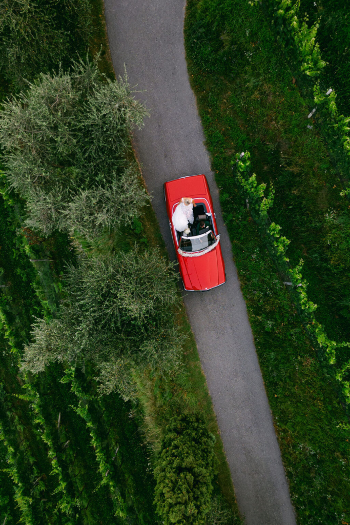 An aerial view captures a red convertible driving along a narrow road bordered by lush green vineyards and trees. A bride in a white gown stands up through the open roof, adding a celebratory feel to the quiet countryside setting.