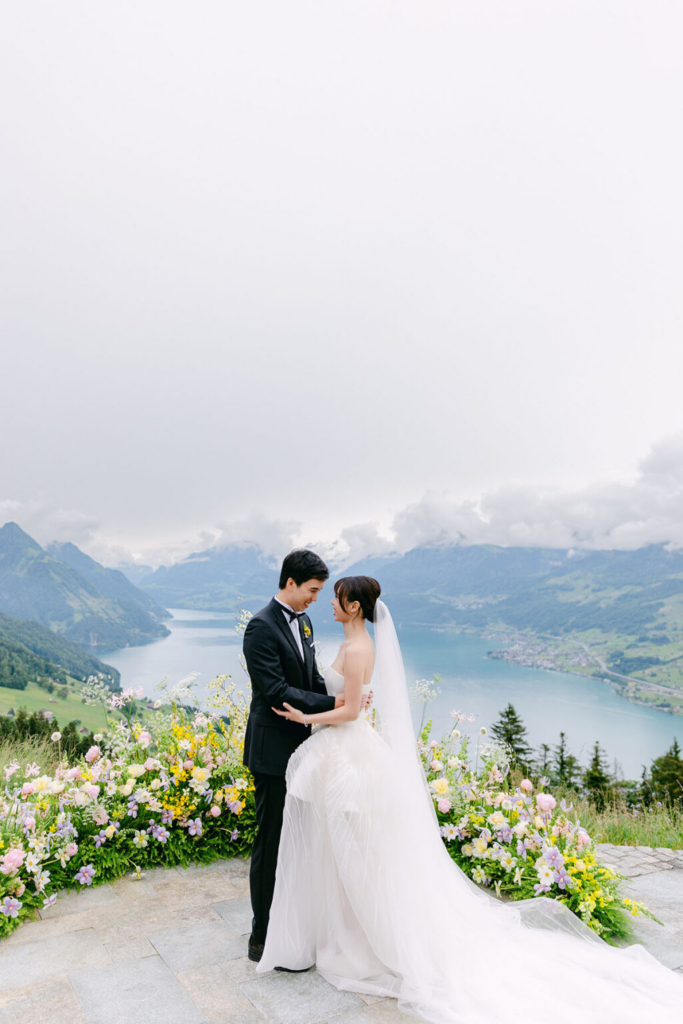 A bride and groom stand together on a mountain overlook framed by pastel-colored flowers, with a lake and rolling green hills stretching behind them. Soft daylight and low clouds create a calm atmosphere as the couple faces each other in an intimate portrait.
