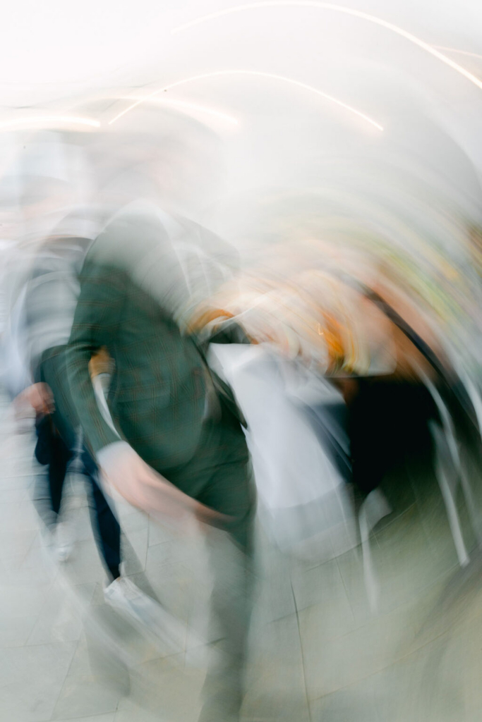 Wedding guests move across a reception space in a photograph created with intentional camera movement and long exposure. The blurred figures, sweeping light trails, and soft colors convey the motion and energy of the celebration while the indoor setting remains faintly visible.