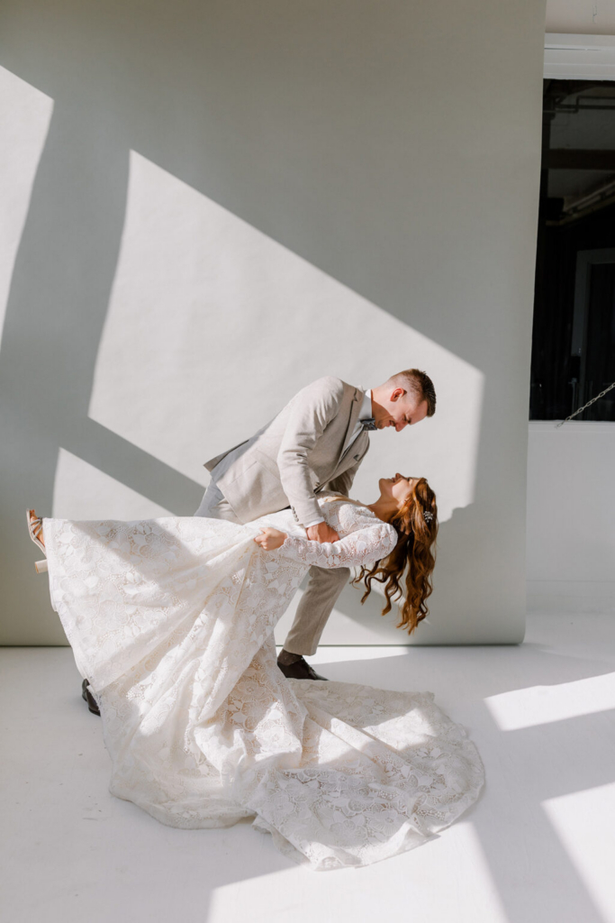 A groom in a light-colored suit dips his bride backward in a bright studio space with white walls and strong geometric shadows. Sunlight streams across the room, highlighting the lace texture of the bride’s gown and the elegant pose of the couple.