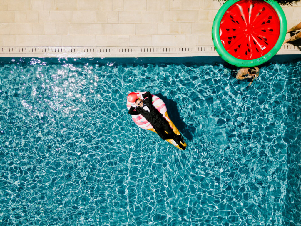 An overhead view shows a man in a black suit and sunglasses floating on a colorful inflatable raft in a bright blue swimming pool. Sunlight reflects across the water, while a large watermelon-shaped float and another swimmer rest near the pool’s edge, creating a playful and relaxed atmosphere