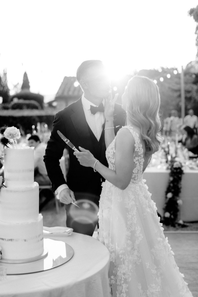 A bride and groom stand beside a multi-tiered wedding cake during an outdoor reception at sunset, photographed in black and white. Backlit by bright sunlight, the bride gently touches the groom’s face while both hold cake knives, surrounded by softly blurred guests and tables.