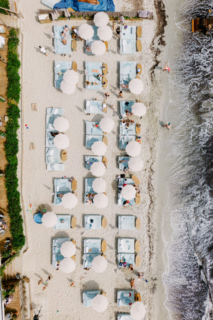 An aerial view looks straight down onto a beach lined with neatly arranged pale-blue lounge beds and white umbrellas beside the shoreline. People relax on the sand and near the water’s edge, while gentle waves roll in from the right side of the frame, creating a bright, summery atmosphere with strong geometric patterns and coastal textures.