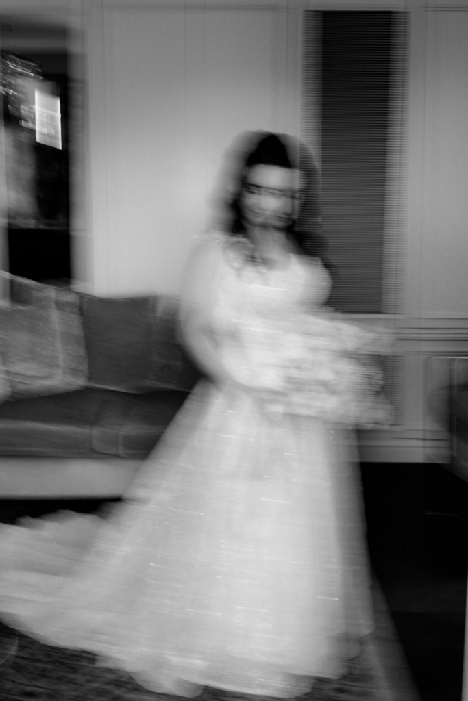 A bride stands indoors holding a bouquet, photographed with intentional camera movement that softens her figure and dress. The black-and-white image emphasizes motion and light within the room while preserving the outline of the bride and her gown.