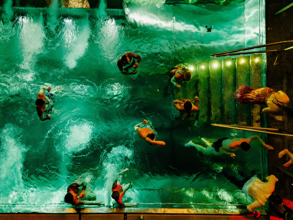 Aerial view of several people gathered in a swimming pool at night, illuminated by green-tinted underwater lighting. Some guests stand on submerged steps while others swim or wade through the water, creating a lively and energetic scene