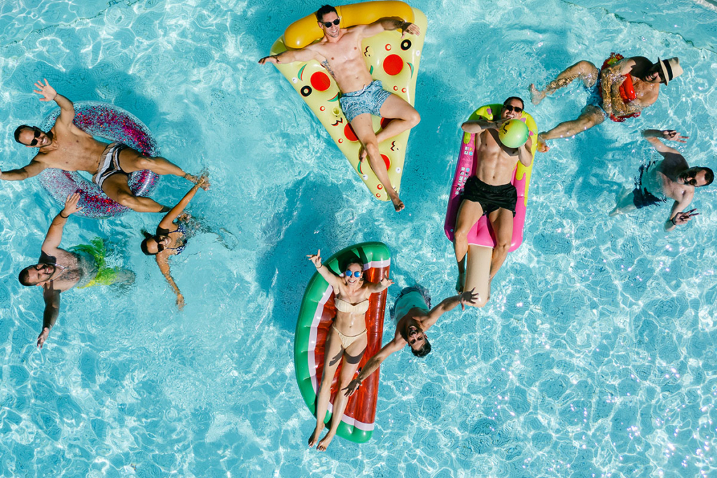 An overhead photograph captures a group of friends enjoying a pool party in bright sunlight, floating on colorful inflatable rafts shaped like pizza slices, popsicles, and watermelon. Guests in the water wave toward the camera, surrounded by sparkling blue water and a cheerful, celebratory mood.