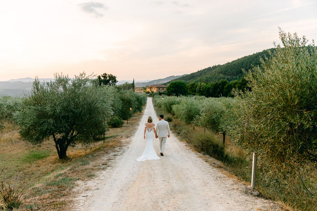 A bride and groom walk hand in hand along a white gravel road lined with olive trees, moving toward a countryside estate in the distance. Soft evening light bathes the landscape, while rolling hills and greenery create a peaceful and expansive setting.