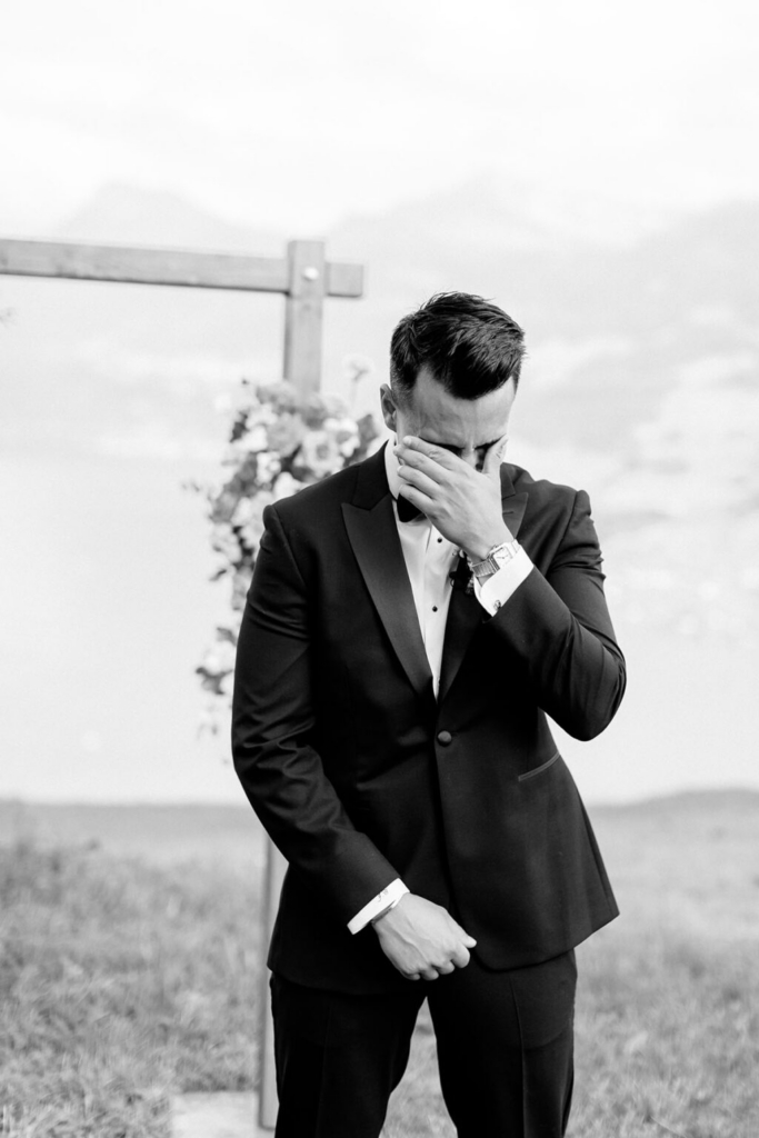 A groom in a black tuxedo stands outdoors during a wedding ceremony, covering his face with one hand while standing in front of a wooden floral arch. The black-and-white image captures a quiet moment against an open landscape, with soft natural light and a calm background.