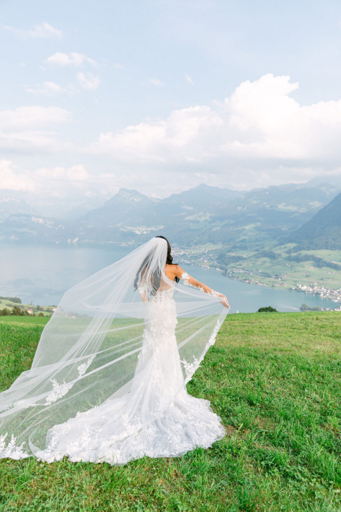 A bride stands on a grassy hillside overlooking a lake and distant mountains, holding out her long veil as it catches the breeze. Soft daylight and layered clouds create an airy, peaceful setting, with the landscape stretching into the distance behind her.