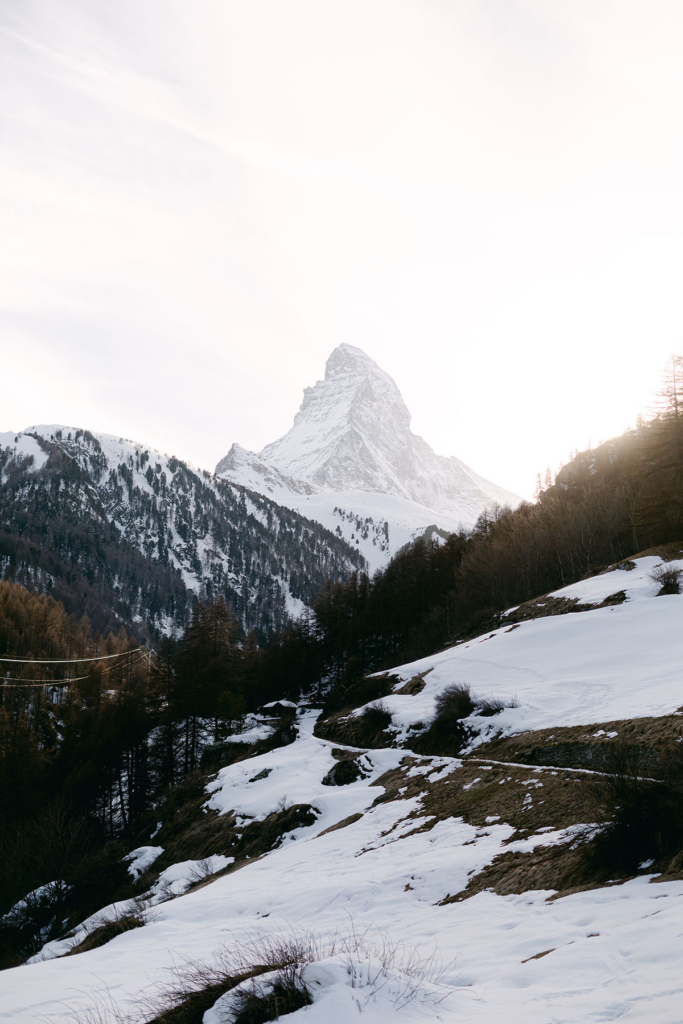 A snow-covered mountain peak rises sharply above a winter landscape of forested slopes and rolling hills. Soft backlight from the low sun brightens the sky behind the summit, while patches of snow and dark evergreens create a calm, expansive alpine scene.