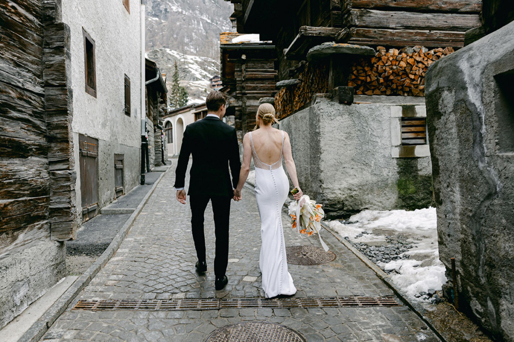 A bride and groom walk hand in hand down a narrow cobblestone street lined with traditional wooden buildings in a mountain village. The bride carries a bouquet of white and orange flowers, while patches of snow and rugged alpine scenery create a quiet, overcast atmosphere.