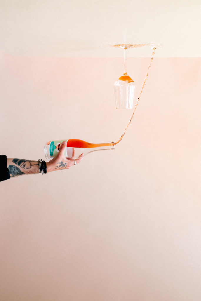 A hand holds a bottle of rosé wine as a stream of liquid pours upward into an upside-down wine glass, creating a playful visual effect. The minimalist composition is set against a soft pastel background, with the motion of the wine frozen in midair.