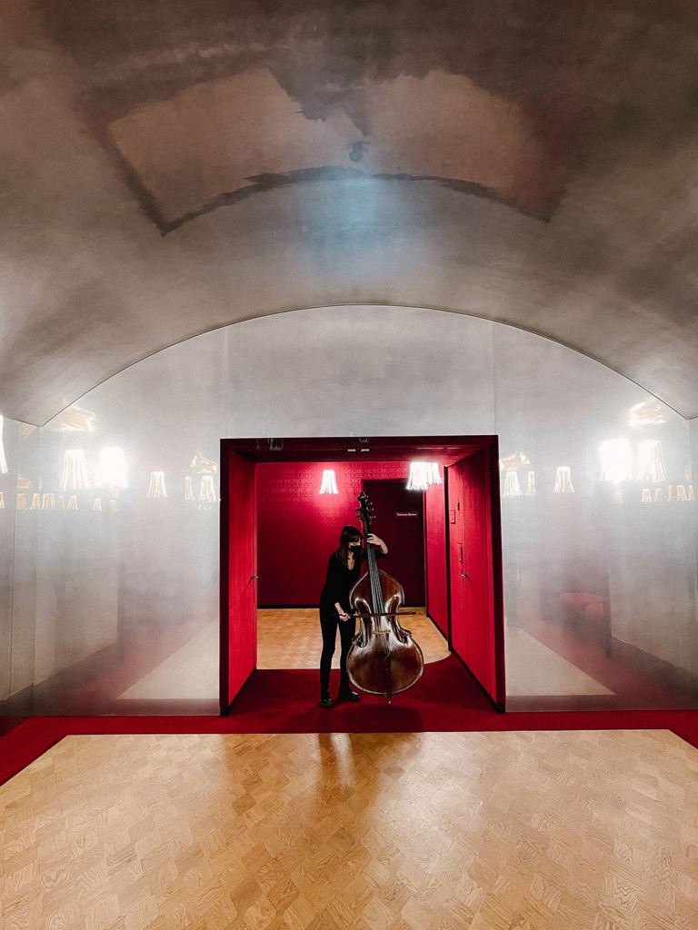 A musician stands in the center of a vaulted hallway playing a double bass, framed by a doorway lined with deep red walls and warm lighting. The polished floor and reflective surfaces create symmetry, while the contrast between the soft gray ceiling and rich red interior gives the scene a dramatic, theatrical mood.